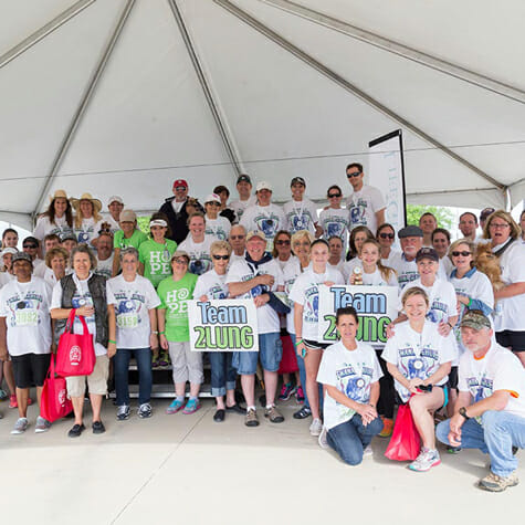 Un gran grupo de asistentes y participantes de la carrera "Donate Life Texas 2nd Chance Run" sonríen para la cámara, de pie en las gradas bajo una gran carpa